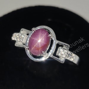 Op de afbeelding: Een zilveren ring met een roze ster-robijn edelsteen in een ronde zetting. De ring heeft kleine diamanten op de band. De ring ligt op een zwarte achtergrond. "Pounak Jewellers" staat in de achtergrond.