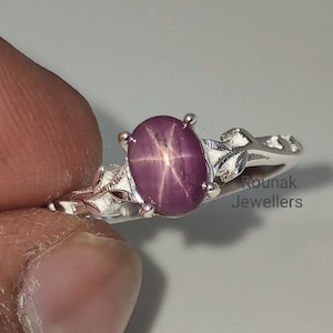 Könnte beinhalten: Ein silberner Ring mit einem rosafarbenen Sternrubin-Edelstein. Der Ring hat ein zartes Blumenmuster und die Worte "Rounak Jewellers" sind in die Band graviert.