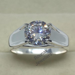 May include: A silver ring featuring a large, round, clear gemstone. The ring is displayed in a white box with the inscription "Rounak Jewellers" on the base.