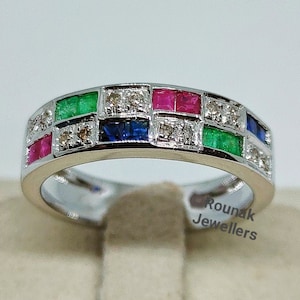 Könnte beinhalten: Ein Silberring mit einer Reihe quadratischer Edelsteine. Die Edelsteine sind rot, grün und blau und in einer weißen Metallfassung gefasst. Der Ring befindet sich auf einem weißen Präsentationsständer.