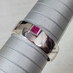 May include: A silver band ring with a square pink gemstone set in the centre. The ring is on a grey display stand. The text "Roupak Jewellers" is visible on the ring.