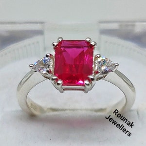 May include: A silver ring featuring a large, square-shaped, bright red gemstone surrounded by three smaller, clear gemstones. The ring is displayed on a white background with the text "Rounak Jewellers".