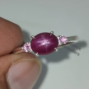 May include: A silver ring featuring a large oval red star ruby gemstone, accompanied by two smaller pink gemstones. The ring is set on a plain band.