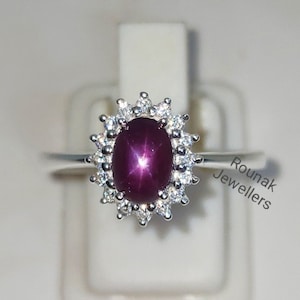 Peut inclure: Une bague en argent avec une grande pierre de rubis ovale entourée d'un halo de petits diamants. Le rubis a un motif en forme d'étoile au centre. La bague est sur un présentoir blanc. Rounak Jewellers est imprimé sur le présentoir.