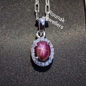 Op de afbeelding: Een zilveren ketting met een hanger met een rode sterrobijn omringd door een halo van kleinere witte diamanten. De hanger is ovaalvormig en de ketting is een delicate schakelstijl. De tekst "Rounak Jewellers" is zichtbaar op de achtergrond.
