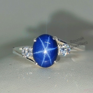 Pode incluir: Um anel de prata com um safira azul estrela e dois diamantes menores. O anel é engastado em uma banda simples e tem um design delicado. Rounak Jewellers.