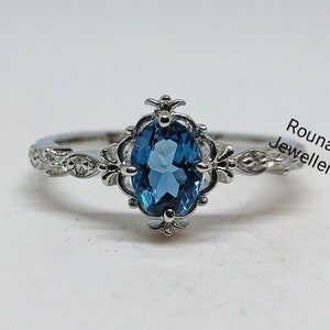 Puede incluir: Un anillo de plata con una piedra ovalada azul engastada en un delicado diseño de filigrana. El anillo está sobre un fondo blanco. 'Rounak Jewellers' está impreso en la esquina superior derecha de la imagen.