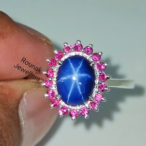 May include: A silver ring featuring a blue star sapphire gemstone encircled by pink rubies. The ring is set in a halo style with a silver band.