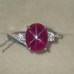 Peut inclure: Une bague en argent avec une grande pierre précieuse en rubis rouge de forme ovale. Le rubis a un motif en forme d'étoile au centre. La bague a deux petites pierres précieuses rondes et blanches de chaque côté du rubis.