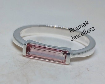 May include: A silver ring featuring a horizontal baguette-cut light pink gemstone set in a smooth metal band. The text Rounak Jewellers is printed in black near the ring. The design showcases a clean, polished finish with a simple setting.