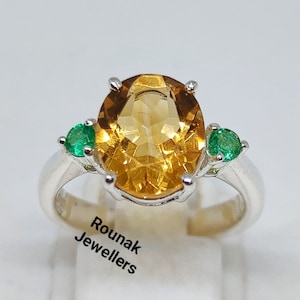 May include: A silver ring featuring a large, oval-cut, golden-yellow citrine gemstone as the centerpiece, flanked by two smaller, round, emerald-green gemstones. The ring is set on a white display stand. The text "Rounak Jewellers" is visible.