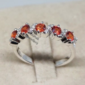 May include: A silver band ring with a row of orange gemstones and smaller white gemstones. The ring is set on a white background.