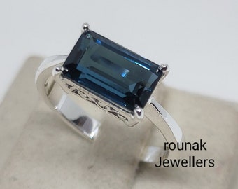 May include: A silver ring featuring a dark blue rectangular gemstone. The ring has a delicate design and is displayed on a white stand. The text "rounak Jewellers" is visible below the ring.