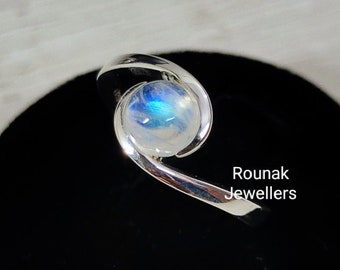May include: A silver ring with a round, iridescent moonstone set in a swirl design. The ring is on a black display stand.  Rounak Jewellers.