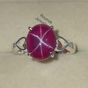 Puede incluir: Un anillo de plata con una gran gema de rubí estrella ovalada. El rubí muestra un patrón de estrella de seis rayos. La banda del anillo tiene recortes en forma de corazón a ambos lados de la gema. El anillo está en una caja de joyería.