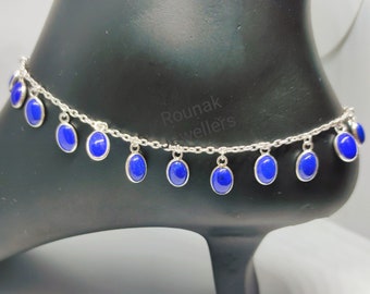 May include: Silver anklet with oval, deep blue gemstones. The gemstones are suspended from a fine chain. The anklet is displayed on a black foot form, highlighting the design. The anklet is a piece of jewellery.
