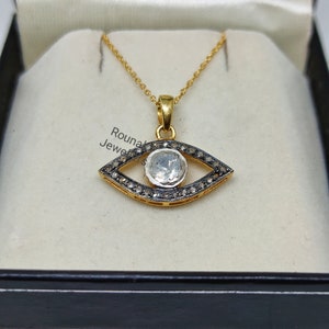 May include: A gold chain necklace with an eye-shaped pendant. The pendant is set with a large, clear gemstone surrounded by smaller black gemstones.