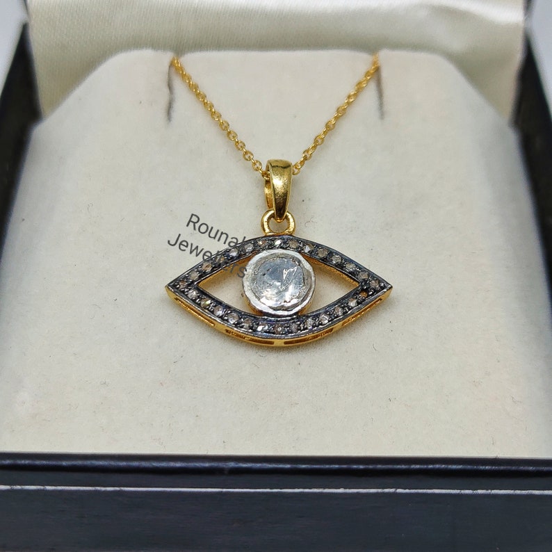 May include: A gold chain necklace with an eye-shaped pendant. The pendant is set with a large, clear gemstone surrounded by smaller black gemstones.