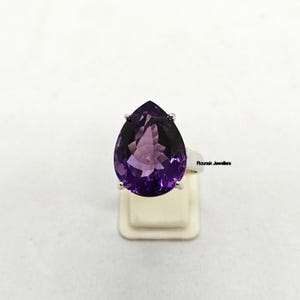 May include: A silver ring featuring a large, pear-shaped, faceted purple amethyst gemstone held by four prongs. The band is smooth and metallic. The text Rounak Jewellers is visible on the side of the ring band near the setting of the stone.