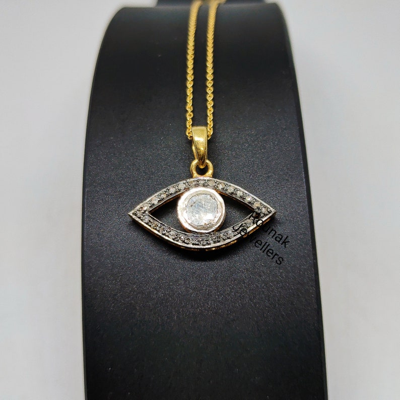 May include: A gold chain necklace with a pendant shaped like an eye. The eye is surrounded by small black diamonds and has a larger white diamond in the center.