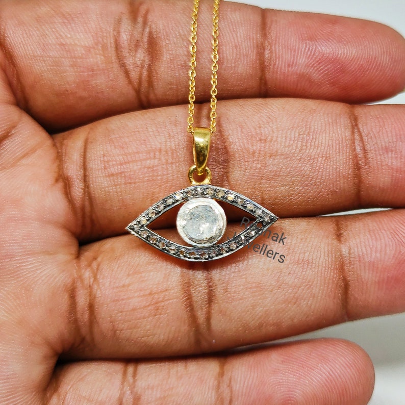 May include: Silver eye pendant necklace with a clear round gemstone in the center, surrounded by smaller clear gemstones. The pendant is on a gold chain.