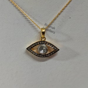 May include: Gold pendant necklace with a large eye-shaped charm featuring a clear gemstone surrounded by smaller black gemstones. The pendant is on a gold chain. The text "Rounak Jewellers" is visible in the background.
