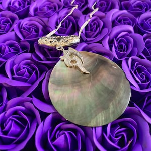 Può includere: Una collana a catena d'argento con un grande ciondolo rotondo, iridescente, in madreperla. Il ciondolo ha un design argentato al centro. La collana è esposta su un letto di rose artificiali viola.