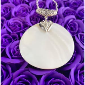 Può includere: Una collana con un ciondolo in madreperla bianca, una catena d'argento e un delicato design a forma di cuore in argento. La collana è esposta su un letto di rose artificiali viola.