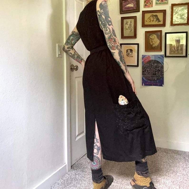 May include: A sleeveless black midi dress featuring a fitted bodice, a defined waistline, and a straight skirt with a side slit. The dress includes a large patch pocket on the right side containing a small, patterned, light-colored object.