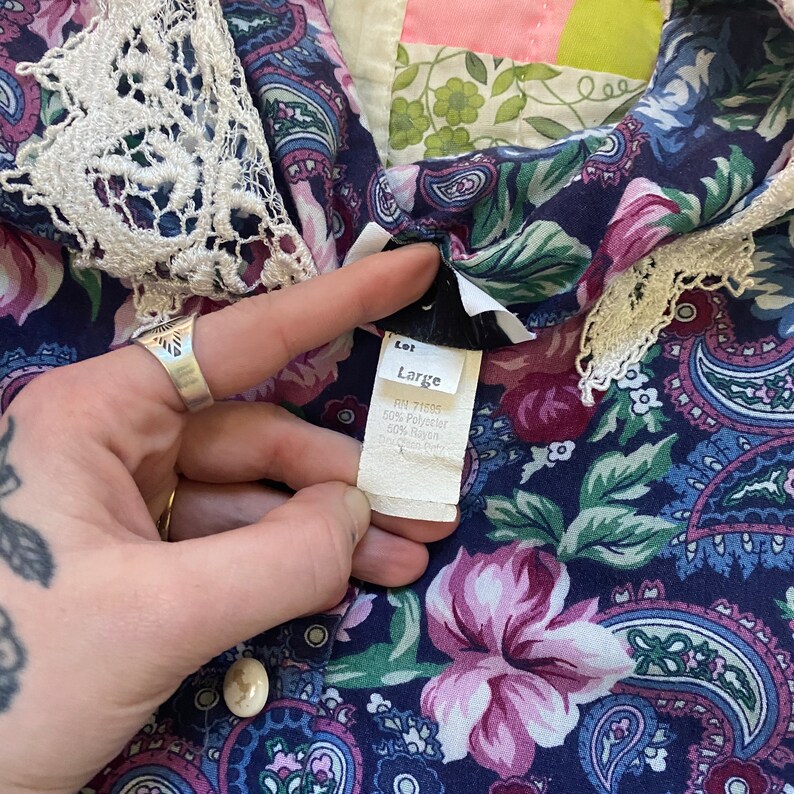 Puede incluir: Una camisa azul oscuro con un patr&oacute;n floral y de cachemira multicolor y un cuello de encaje color crema. Se ve una etiqueta de tela blanca con el texto "Lot Large", "RN 71605", "50% Polyester" y "50% Rayon" impreso en color negro.