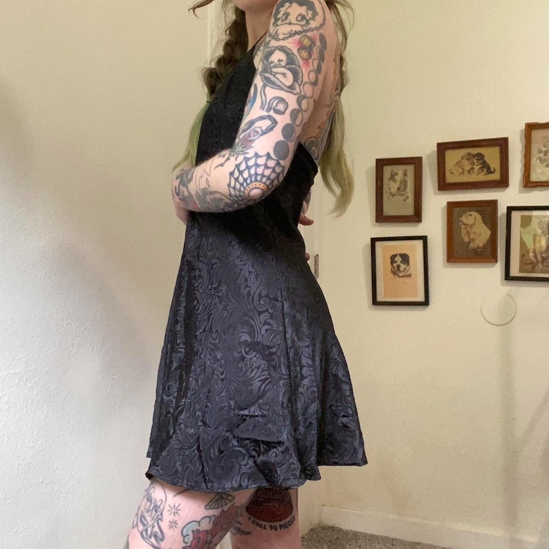 May include: A black satin mini dress with a floral pattern. The dress has a halter neckline and a fitted bodice. The model is wearing the dress with a black bra.