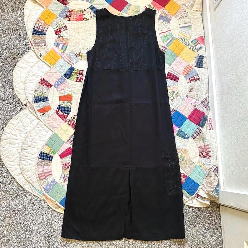 May include: A sleeveless black midi dress featuring a solid black lower section and a textured black upper bodice. The garment includes a subtle tonal embroidered floral pattern on the lower right side and a vertical center slit at the hemline.