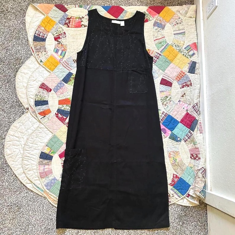 May include: A black sleeveless midi dress featuring a round neckline, a textured upper bodice panel, a small chest pocket, and a larger patch pocket on the lower left side. The garment is constructed from a solid black fabric with a consistent tone.