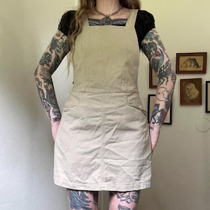 May include: A tan overall dress with a square neckline and two front pockets. The dress is made of a sturdy fabric and has a simple, classic design. The dress is a versatile piece that can be dressed up or down.