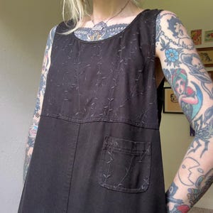 May include: A black sleeveless dress featuring a round neckline and a single patch pocket on the front. The upper bodice and the pocket are decorated with tonal black embroidered vine patterns that extend across the chest and pocket area.