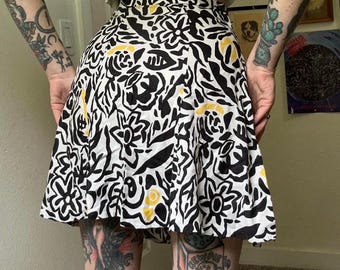 May include: A high-waisted skirt featuring a bold, abstract black and white floral pattern with scattered yellow accents. The skirt has a flared, A-line silhouette that falls above the knee, constructed with a fitted waistband and a smooth, lightweight fabric.