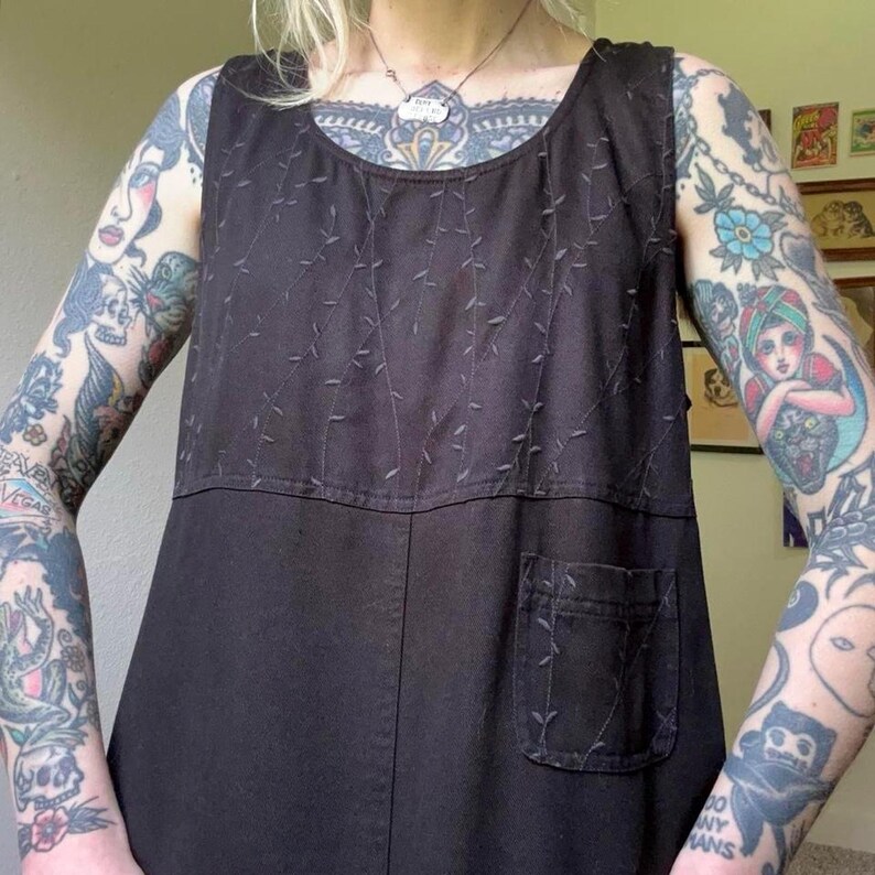 May include: A sleeveless black tunic featuring a round neckline and a single patch pocket on the lower front. The upper portion of the garment is decorated with a subtle, tonal embroidered vine pattern that extends down to the pocket.