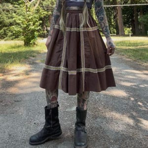 May include: A brown midi skirt featuring a high-waisted, pleated design with a grid pattern formed by white and yellow zigzag trim. The skirt has a structured waistband and falls to mid-calf length, paired with black leather mid-calf boots.