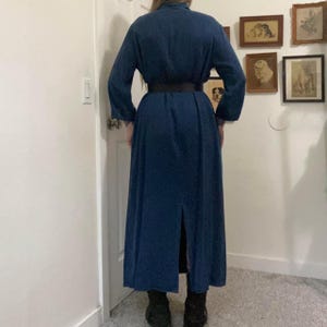 Puede incluir: Un vestido largo de color azul oscuro con cuello, una abertura en la espalda y un cintur&oacute;n negro. El vestido tiene mangas largas y un corte holgado.