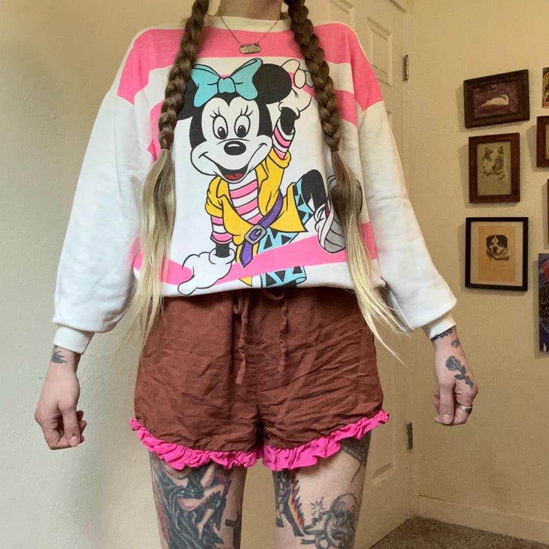 May include: A white sweatshirt with pink stripes and a graphic of Minnie Mouse. The shorts are brown with a pink ruffle trim. The outfit is casual and vintage.