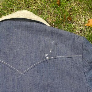 May include: A dark blue denim jacket with a white sherpa collar. The jacket has a white stitching detail around the collar and pockets. There are a few light-colored marks on the jacket.