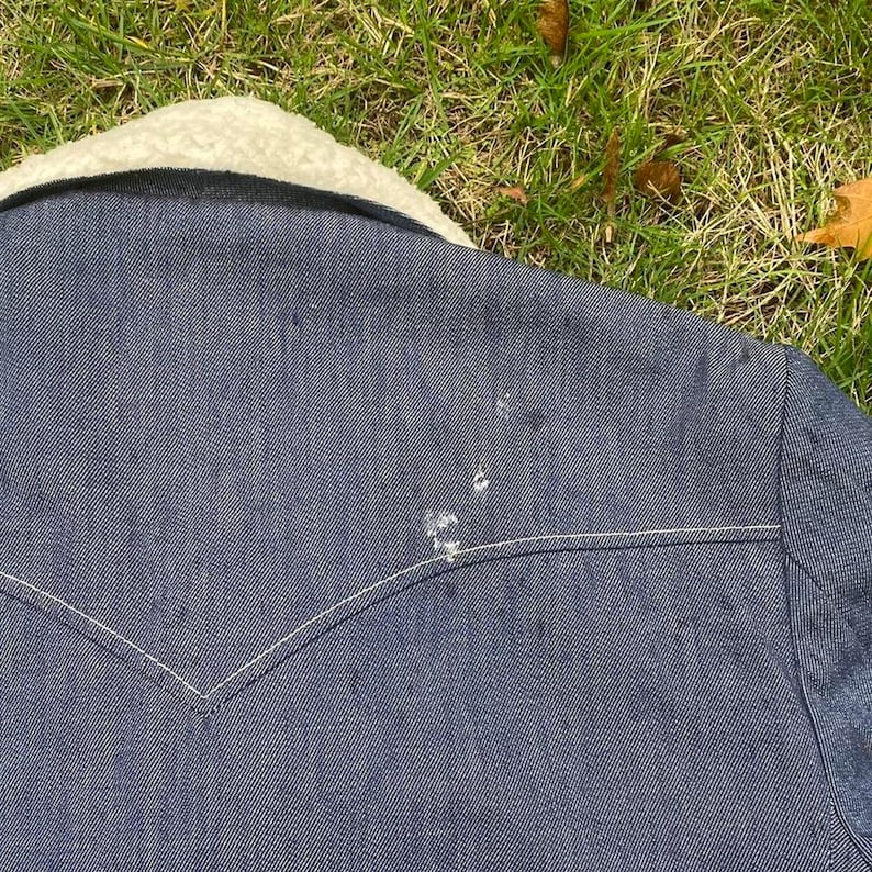 May include: A dark blue denim jacket with a white sherpa collar. The jacket has a white stitching detail around the collar and pockets. There are a few light-colored marks on the jacket.