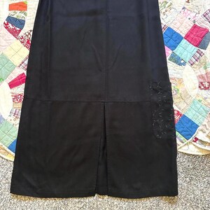 May include: A solid black skirt featuring a straight silhouette, a horizontal seam across the lower portion, a central vertical slit at the hem, and a single patch pocket on the right side adorned with a subtle, tonal embroidered leaf pattern.
