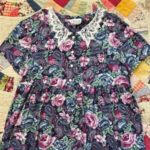 Puede incluir: Un vestido de manga corta con fondo azul oscuro y un estampado floral y de cachemira multicolor. La prenda incluye un cuello de encaje blanco, una tapeta frontal con cinco botones blancos y una etiqueta que dice LARISA NEW YORK.
