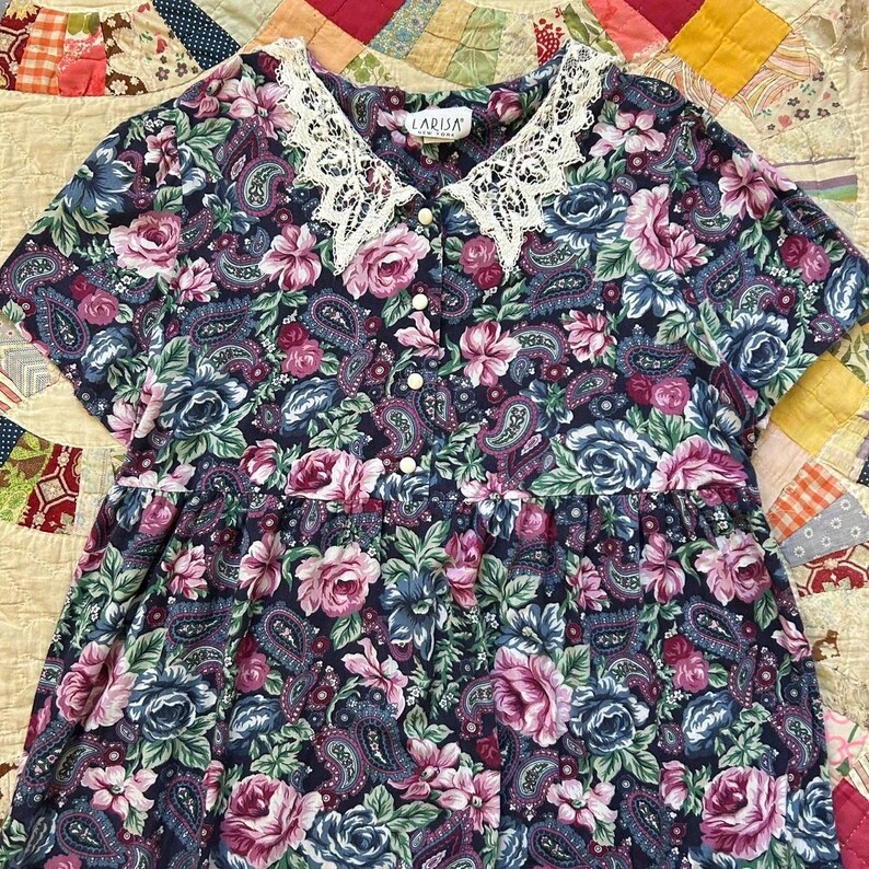 Puede incluir: Un vestido de manga corta con fondo azul oscuro y un estampado floral y de cachemira multicolor. La prenda incluye un cuello de encaje blanco, una tapeta frontal con cinco botones blancos y una etiqueta que dice LARISA NEW YORK.