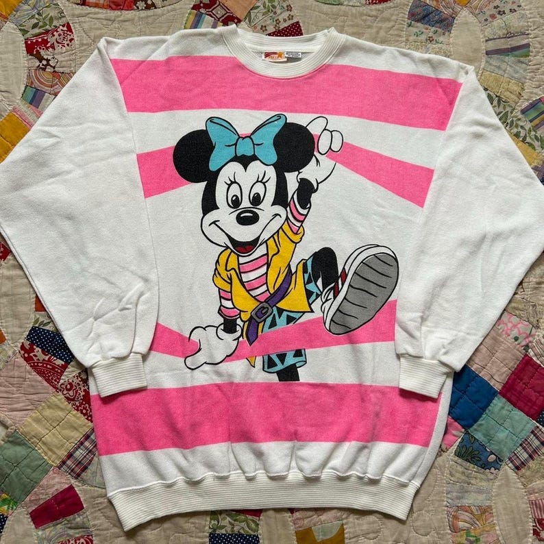 May include: White sweatshirt with pink horizontal stripes and a Minnie Mouse graphic. Minnie is wearing a yellow jacket, blue bow, and a colorful outfit. The sleeves are white.