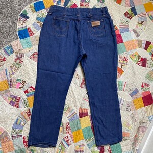 Puede incluir: Un par de pantalones vaqueros de mezclilla azul oscuro con un corte recto cl&aacute;sico. Los pantalones tienen un parche de cuero en la parte posterior con el logotipo de Wrangler.