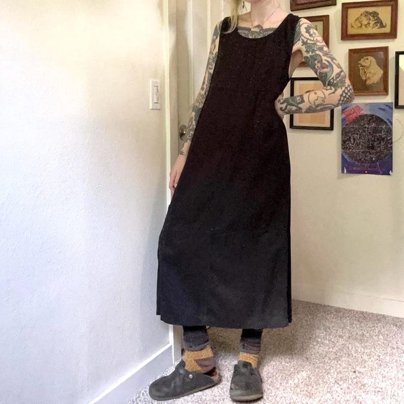 May include: A sleeveless black midi dress with a scoop neckline and a straight silhouette. The dress features a solid dark fabric that falls below the knees, paired with dark leggings, mustard-colored socks, and dark gray slip-on clogs.