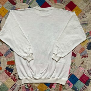 May include: A white crew neck sweatshirt with long sleeves and ribbed cuffs and hem. The sweatshirt is made of a soft, comfortable material. The back of the sweatshirt is visible, and it is laying on a colorful patchwork quilt.