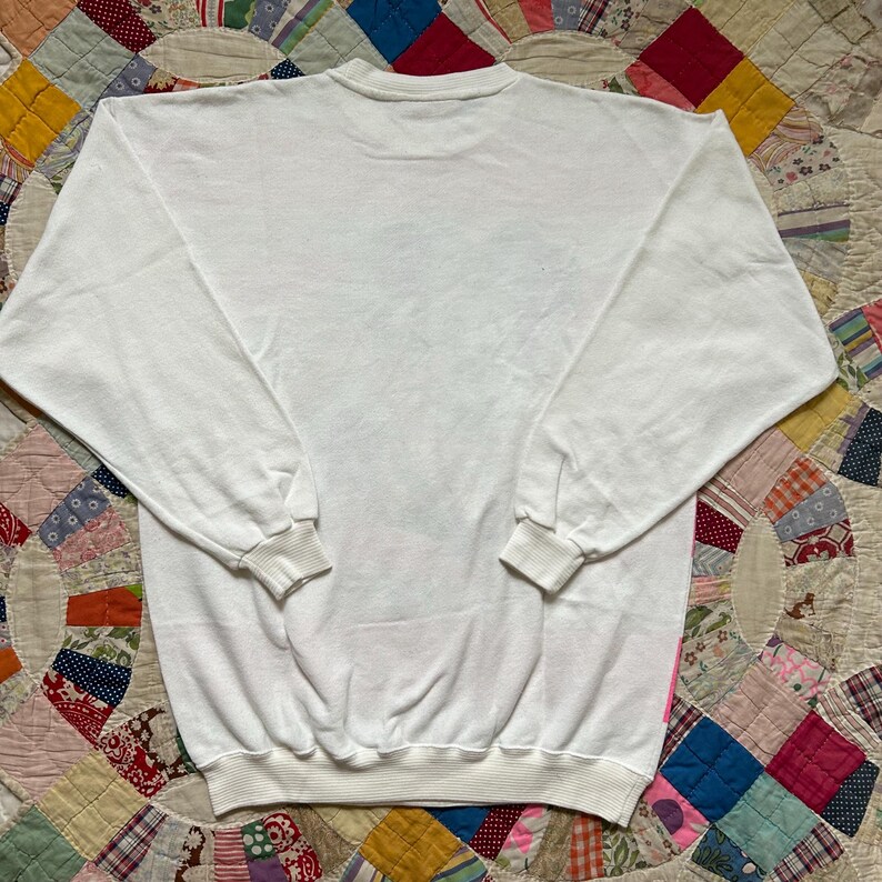 May include: A white crew neck sweatshirt with long sleeves and ribbed cuffs and hem. The sweatshirt is made of a soft, comfortable material. The back of the sweatshirt is visible, and it is laying on a colorful patchwork quilt.